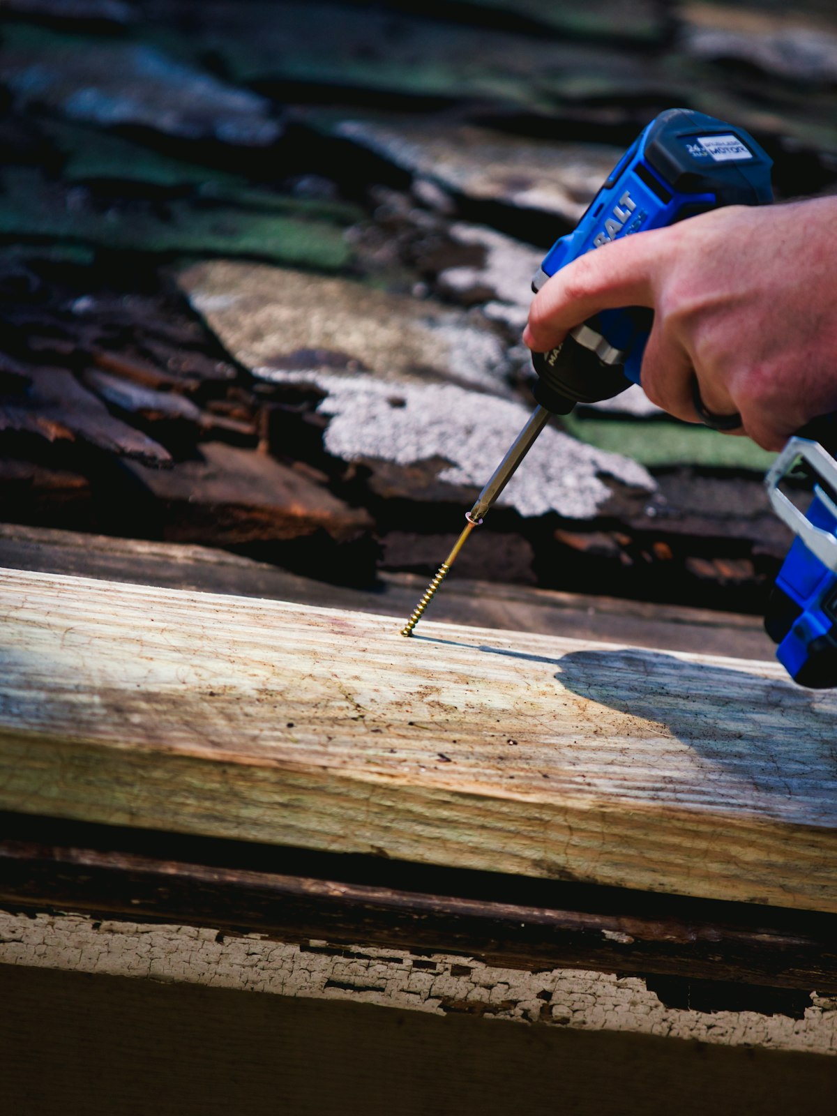 A gloved hand driving a screw into a roof timber with a cordless drill